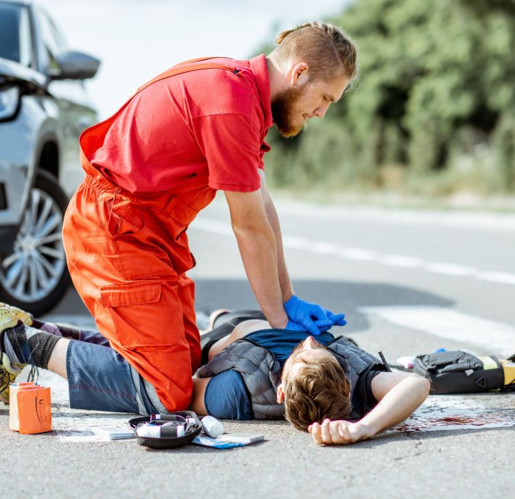 A man in a red jumpsuit performs CPR on another man lying on the road. An open first aid kit is nearby, and a vehicle is parked in the background. The scene appears to be an emergency situation.