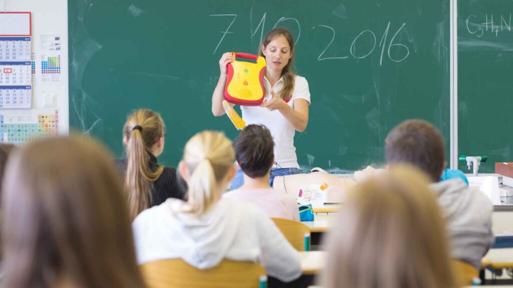 A teacher stands in front of a classroom holding a defibrillator and addressing students. The students, seated and facing the teacher, listen attentively. A chalkboard with writing is behind the teacher in the classroom setting.