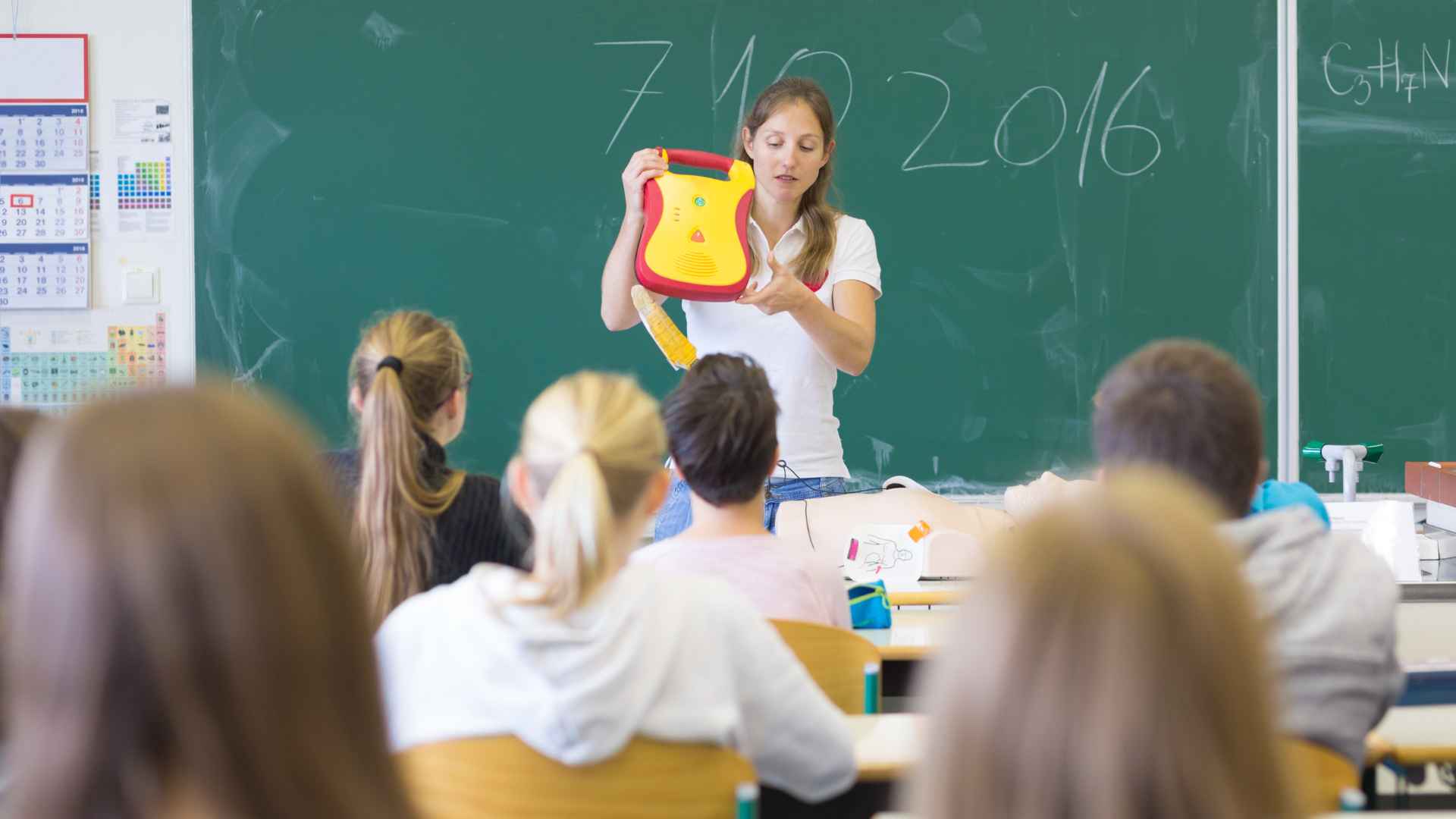 A teacher stands in front of a classroom holding a defibrillator and addressing students. The students, seated and facing the teacher, listen attentively. A chalkboard with writing is behind the teacher in the classroom setting.