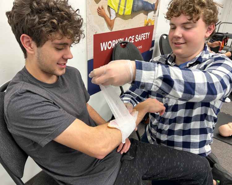 Two young men in a classroom setting practice first aid. One is seated, extending his arm while the other, wearing gloves, applies a bandage to the forearm. A safety poster is visible in the background.