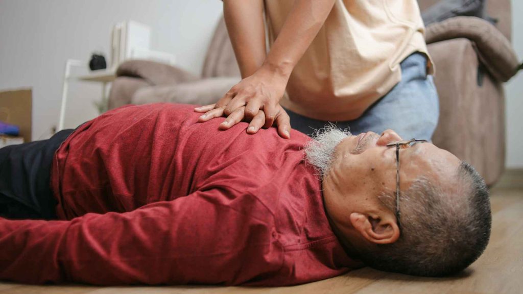 close up of an older man in a red long sleeved shirt laying on the floor receiving CPR from a woman