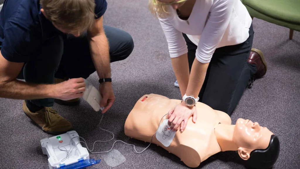 a man and woman practicing cpr and using an aed in a cpr training course. the woman is performing compressions on the manikin while the man is placing the aed pads on it.