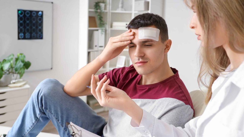 young man at doctor's office holding bandaged head talking to doctor