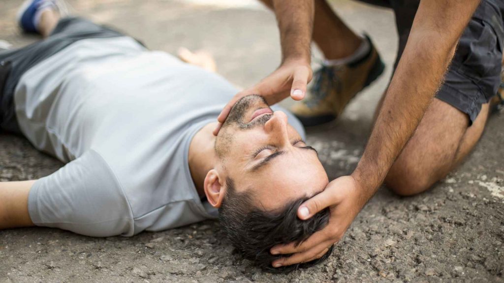 unconscious man laying on his back on the ground. another person is performing a head-tilt, chin-lift to open his airway