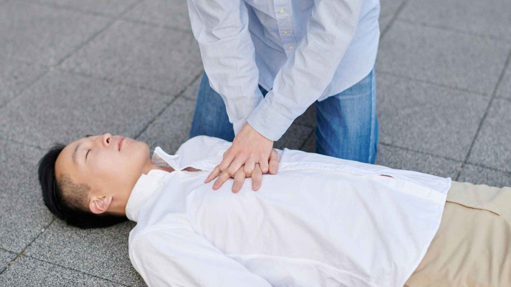 man laying on his back unconscious on the ground. another man is performing chest compressions on him.