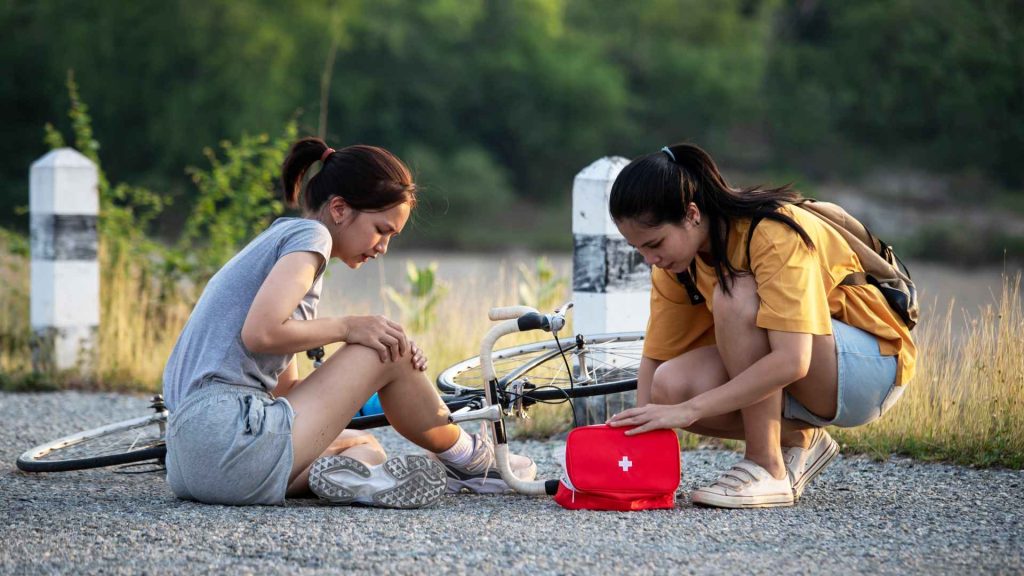 girl opening up first aid kit to assist another girl who is holding her knee with a bike on it's on in the background.