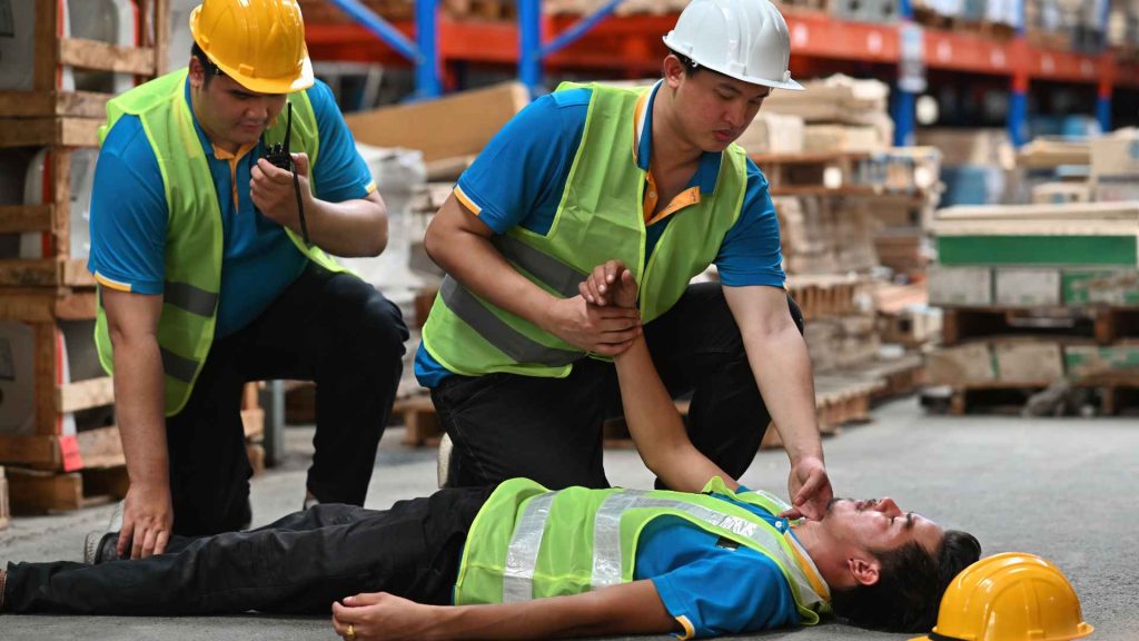 two warehouse workers checking on a coworker who is laying on the ground, potentially unconscious