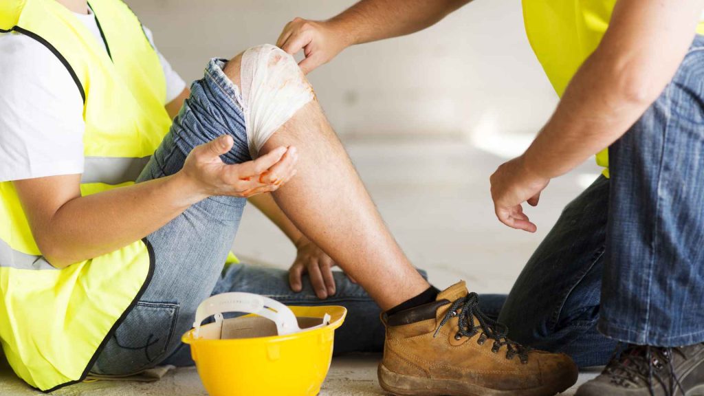 a construction worker sitting with his knees up while another construction worker bandages the knee of the one sitting down