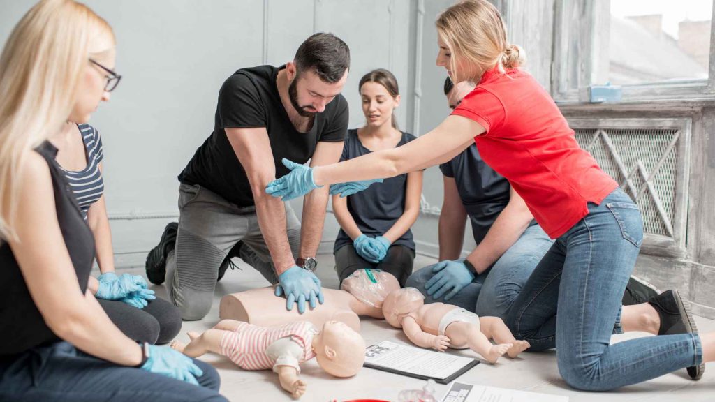 a first aid and cpr training class of men and women. a man is practicing cpr on a manikin and a female instructor is guiding him