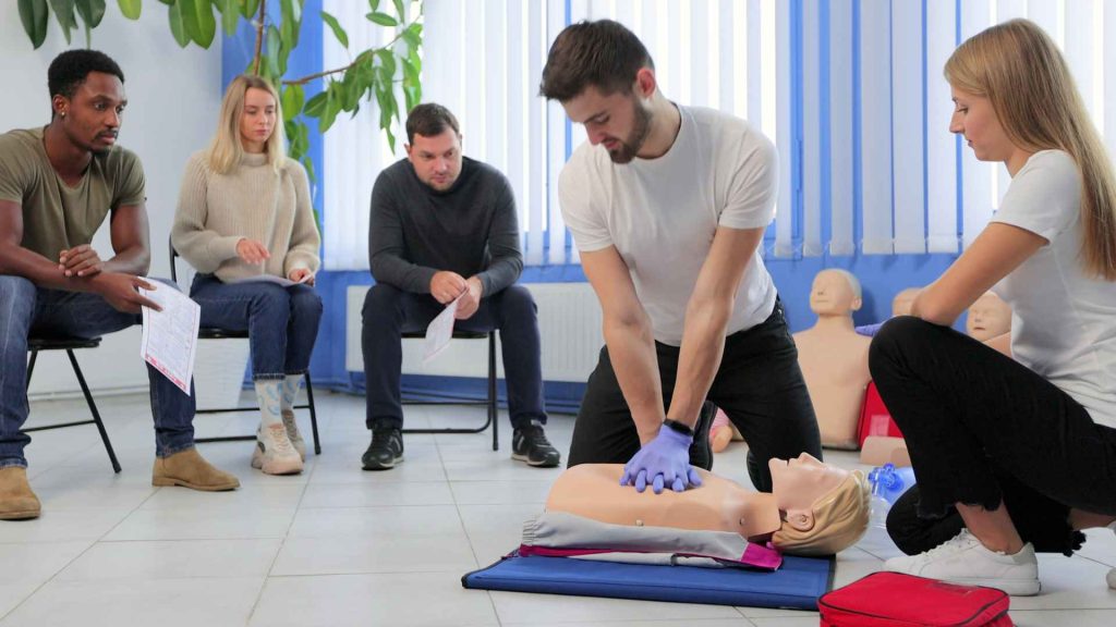 man practicing cpr on a manikin with additional students sitting in the background on chairs