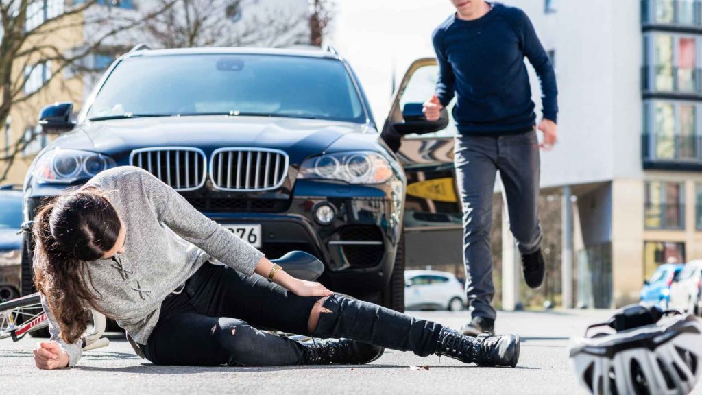 a male driver rushing over to a woman he hit with his car. the woman is sitting on the ground holding her leg