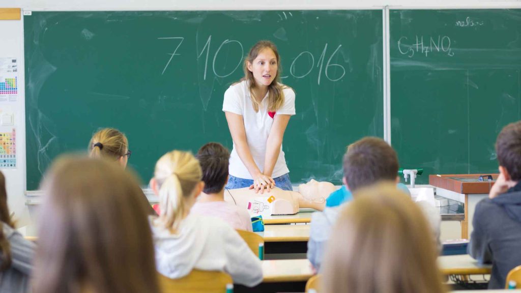 a teacher displaying how to perform cpr on a manikin in front of a class of teenagers
