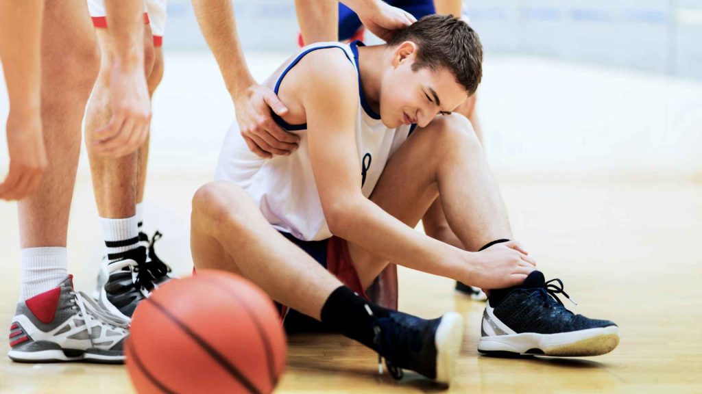 young man in basketball uniform sitting on the court holding his ankle in pain. his teammates are checking if he is ok