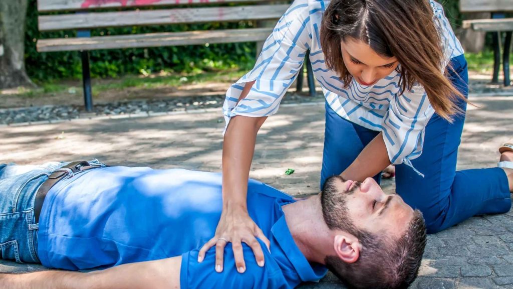 woman kneeling next to a man who is laying on the ground outside with his eyes clothes. she is concerned, with her hands on his shoulders to see if he's ok