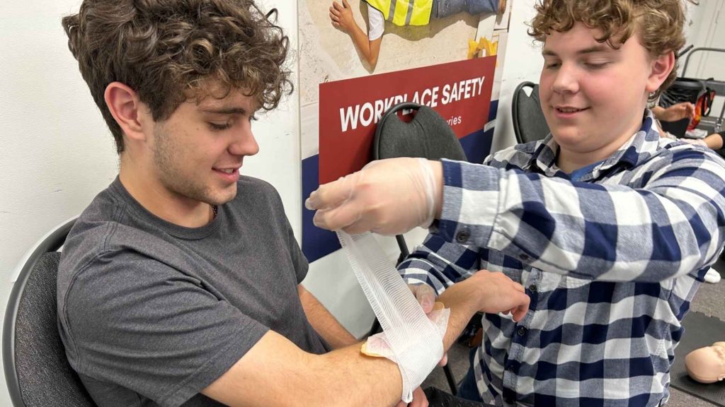 two male students in a first aid training room practicing life-saving skills. the male on the right is practicing applying a bandage to the male on the left's arm.