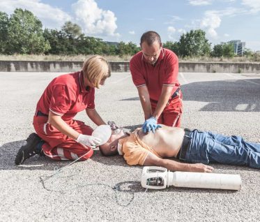 Two rescuers in red uniforms perform CPR on a man lying on the ground. The man is shirtless with an oxygen mask placed by one rescuer, while the other performs chest compressions. An oxygen tank is beside them on the pavement.