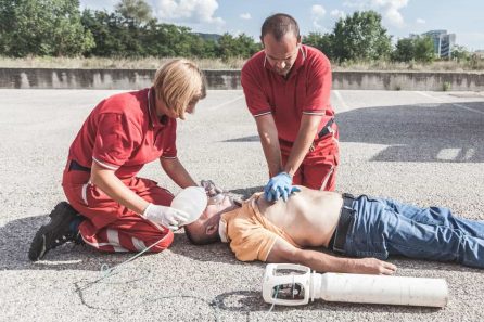 Two rescuers in red uniforms perform CPR on a man lying on the ground. The man is shirtless with an oxygen mask placed by one rescuer, while the other performs chest compressions. An oxygen tank is beside them on the pavement.
